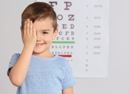 Young boy covering one eye while smiling in front of an eye chart.
