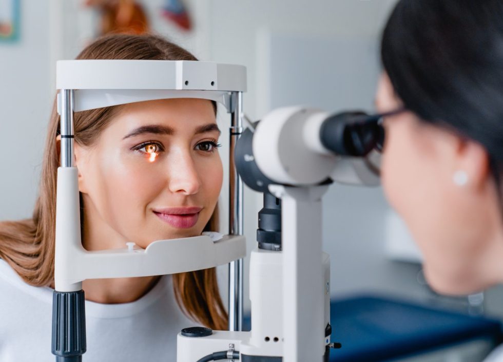 Woman undergoing an eye examination with a slit lamp while an optometrist observes.
